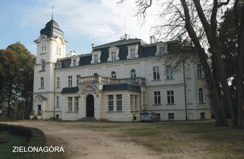 Pałac w Zielonejgórze widziany w dzień. Biały budynek z jedną wieżyczką, otoczony drzewami, do którego prowadzi ścieżka. Przed budynkiem zaparkowany samochód.
